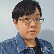 An image of Emery Ly. She is a light skinned Chinese person with short black hair and brown eyes. She wears a pair of glasses and a dark shirt underneath a denim vest.
